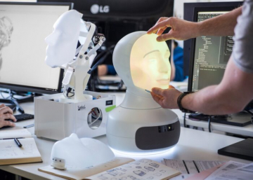 A person fine-tunes a the Furhat robot head with a projected face in a tech lab setting.