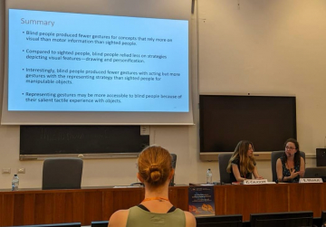 In the picture, you can see Ezgi Mamus standing to the right of a large digital screen displaying a presentation with the title "How do people describe the world without sight?". Ezgi, wearing a red sweater and black jeans, is gesturing with her hands as she speaks.
