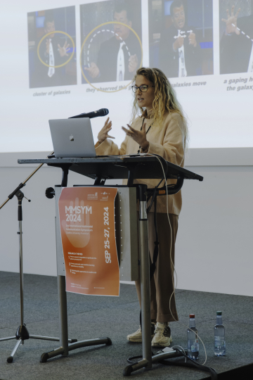 A woman in glasses wears a beige sweater, loose brown pants, and white sneakers while presenting at a conference podium.