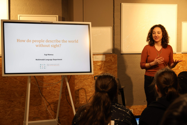 In the picture, you can see Ezgi Mamus standing to the right of a large digital screen displaying a presentation with the title "How do people describe the world without sight?". Ezgi , wearing a red sweater and black jeans, is gesturing with her hands as she speaks.