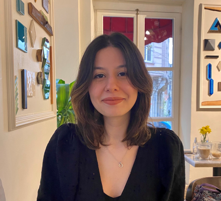 A woman with shoulder-length dark brown hair smiles softly in a cozy café, seated near colorful wall art and a window.