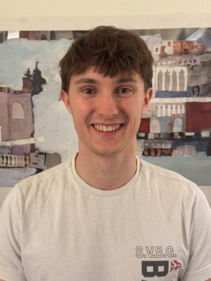 A young man with brown hair smiling at the camera, wearing a white T-shirt, standing in front of abstract artwork.